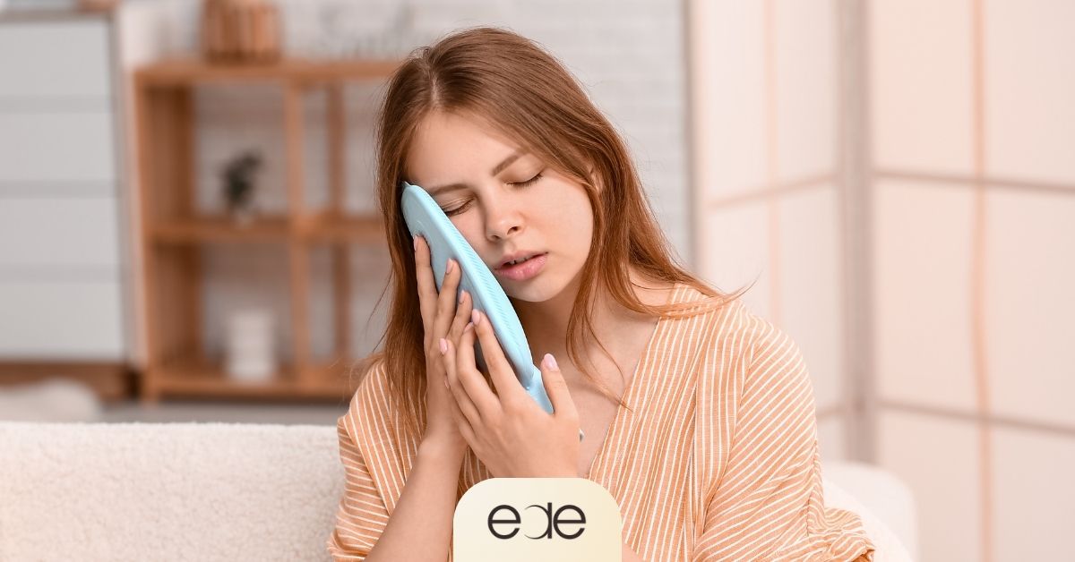 A young woman holding a blue cold compress to her cheek to reduce pain and swelling during recovery after a tooth extraction.
